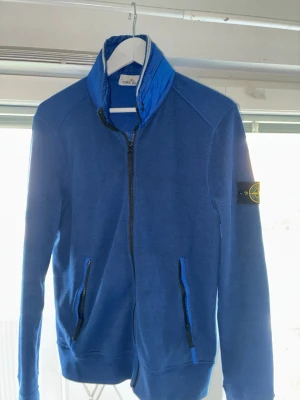 Stone Island jacka/kofta - Blå kofta/jacka från Stone Island med dunkrage och svarta dragkedjor. Tröjan har två fickor framtill och den klassiska Stone Island-loggan på vänster ärm. Perfekt för vår/sommarkvällar