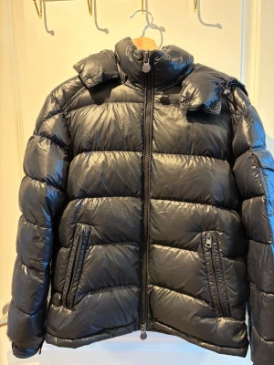 Svart pufferjacka från Moncler - Säljer en svart pufferjacka från Moncler med glansig finish och hög krage. Jackan har två snedställda fickor med dragkedja och en kraftig dragkedja framtill. Klassisk dunjacka med quiltad design och diskret logga på dragkedjan.