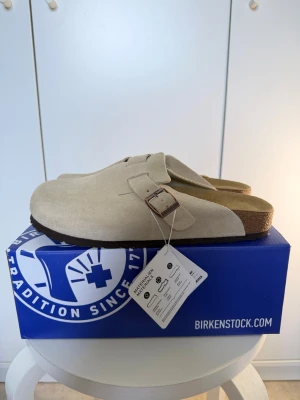 Birkenstock Boston tofflor khaki - Säljer ett par Birkenstock Boston tofflor i färgen khaki. De har ovandel i mocka, justerbart spänne i metall och klassisk korksula med mjuk fotbädd. Perfekta för dig som gillar stilren och bekväm design.