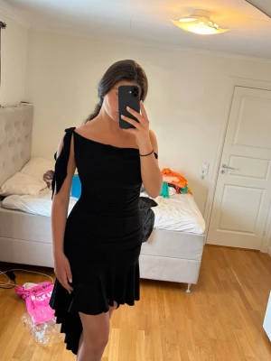 Svart oneshoulder klänning med volang - Säljer en svart oneshoulder klänning med volangkant och asymmetrisk design. Klänningen har en snygg knyt-detalj på axeln och sitter figurnära upptill. Perfekt för dig som vill sticka ut med en unik och trendig look.