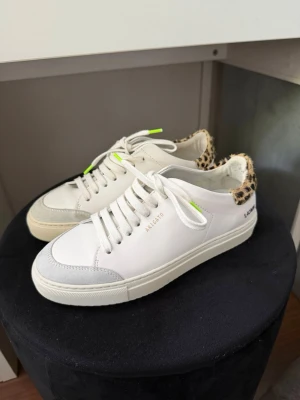Axel Arigato vita sneakers med leoparddetalj - Snygga vita sneakers från Axel Arigato med grå mockadetaljer på tån och cool leopardmönstrad häl. Skorna har vita snören och små neonfärgade detaljer. Perfekta för dig som vill sticka ut med en unik touch på klassiska sneakers. Endast använda två gånger!