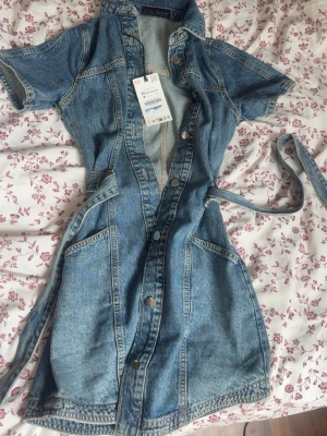 Kort jeansklänning med knappar - Snygg kort jeansklänning från Zara  i blå denim med knappar framtill och tillhörande bälte i midjan. Klänningen har korta ärmar, krage och stora fickor framtill. Perfekt för dig som gillar en avslappnad men trendig stil.