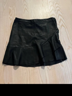 Svart skinnkjol med volang - Snygg svart kortkjol i skinn med volangdetalj längs nederkanten. Kjolen har en lätt asymmetrisk skärning och ger en cool vibe till din outfit. Perfekt att matcha med en enkel topp eller stickad tröja.