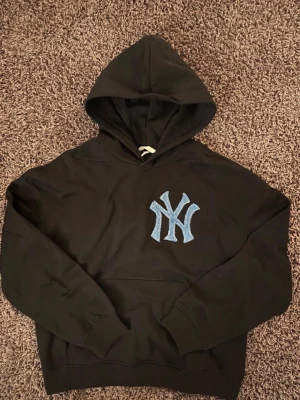Svart hoodie med NY-logga i denim - Svart hoodie med stor huva och en blå denim NY-logga på bröstet. Hoodien har klassisk känguruficka och ribbade muddar vid ärmslut och nederkant. Perfekt för dig som gillar streetwear och sportiga vibes. Handsytt märke 