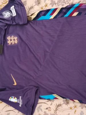 Lila England fotbollströja Nike - Säljer en lila England fotbollströja från Nike med guldfärgad swoosh och England-emblem på bröstet. Tröjan har UEFA EURO 2024-märke på ärmen och färgglada detaljer längs sidan. Perfekt för dig som älskar fotboll och vill sticka ut på läktaren.
