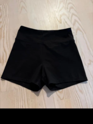 Svarta högmidjade träningsshorts - Säljer ett par svarta högmidjade shorts i stretchigt material, perfekta för träning eller chill. De har en clean look utan synliga detaljer och sitter tajt mot kroppen för en snygg siluett.