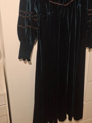 Mörkgrön sammetsklänning med detaljer - Mörkgrön långklänning i mjuk sammet från H&M. Klänningen har långa puffärmar med smock vid ärmslut och dekorativa band i bronsfärg över byst och ärmar. Rund halsringning och elegant fall. Perfekt för dig som gillar vintagevibbar och unika detaljer.
