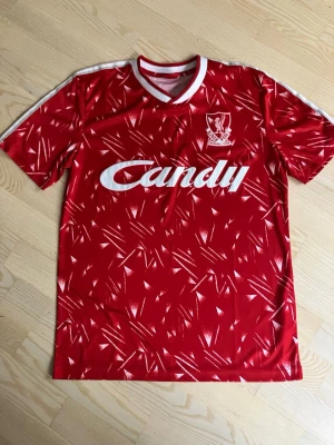 Liverpool retro matchtröja röd - Säljer en klassisk röd Liverpool retro matchtröja med vitt Candy-tryck på bröstet och klubbmärke på vänster sida. Tröjan har ett mönster i vitt och rött, rund hals med vit och röd kant samt vita detaljer på axlarna. Perfekt för dig som älskar fotboll och vintage stil.