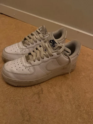 Nike Air Force 1 vita sneakers - Klassiska vita Nike Air Force 1 sneakers med lågt skaft. Skorna har perforerad tå, vita skosnören och Nike-logga på sidan samt på plösen. Tillverkade i läder med gummisula och diskreta detaljer i vitt. Perfekta för en clean och tidlös stil.