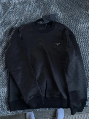 Svart sweatshirt från Prada - Snygg svart sweatshirt från Prada med klassisk rund halsringning och ribbade muddar. Tröjan har en diskret ton-i-ton struktur och det ikoniska metalltriangel-logotypen på bröstet. Perfekt för en stilren och exklusiv look. Vid frågor kontakta mig via dm svara så snabbt jag kan