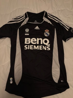 Real Madrid svart matchtröja Adidas S - Svart Real Madrid matchtröja från Adidas med vita detaljer och BenQ Siemens-tryck på bröstet. Tröjan har klubbmärke, Adidas-logga och klassiska ränder på axlarna. Tillverkad i lätt och ventilerande material, perfekt för fotboll eller träning.