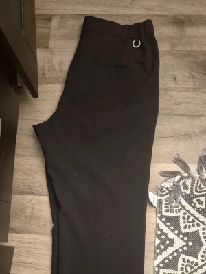 Svarta kostymbyxor från Stockh LM - Svarta kostymbyxor från Stockh LM med rak passform och diskreta fickor. Byxorna har hällor i midjan och stängs med dragkedja, knapp och hake. Snygg och stilren design som passar till många olika looks.