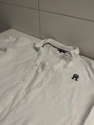 Vit henley-skjorta från Tommy Hilfiger - Vit henley-skjorta från Tommy Hilfiger med korta ärmar och knappar framtill. Skjortan har ett broderat TH-logotyp på bröstet och är tillverkad i mjuk bomull. Perfekt för en clean och stilren look.
