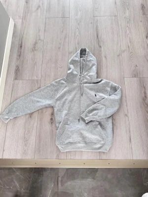 Grå hoodie från Polo Ralph Lauren - Snygg grå hoodie från Polo Ralph Lauren med klassisk huva och dragsko. Har en broderad logga på bröstet och en stor magficka framtill. Tillverkad i mjukt material som är skönt att ha på sig. Perfekt för en avslappnad och stilren look.