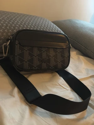Svart axelväska Lacoste geometriskt mönster - Svart axelväska från Lacoste med geometriskt mönster i vitt och bred justerbar axelrem. Väskan har dragkedja upptill, en ficka framtill och detaljer i konstläder. Perfekt för dig som vill ha en stilren och modern look.