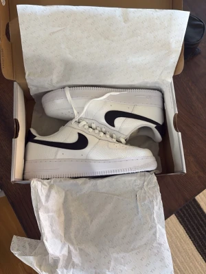 Nike Air Force 1 vita sneakers - Klassiska Nike Air Force 1 sneakers i vitt läder med svart swoosh på sidan. Lågt skaft, perforerad tåbox för ventilation och vit sula. Tidlös design som passar till det mesta och ger en clean look. Sköna att gå i och riktigt snygga till jeans eller shorts.