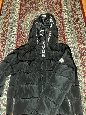 Svart dunjacka från Moncler med huva - Snygg svart dunjacka från Moncler med stor huva och logotyp på ärmen. Jackan har dragkedjor på fickorna och Moncler-tryck på huvan. Perfekt för kalla vinterdagar och riktigt trendig look.