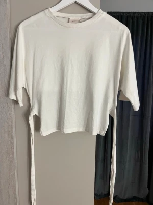 Vit topp med knytband - Trendig vit topp med korta ärmar och långa knytband i sidorna som kan knytas i ryggen för en snygg detalj. Tillverkad i mjuk bomull med rund halsringning. Perfekt för att skapa en avslappnad men ändå stilren look. Knappt använd och inga defekter!💕