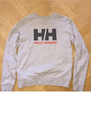 Grå sweatshirt från Helly Hansen - Snygg grå sweatshirt från Helly Hansen med stor svart HH-logga och röd text på bröstet. Tröjan har rund halsringning, långa ärmar och ribbade muddar vid ärmslut och nederkant. Perfekt för en avslappnad och sportig stil.