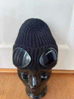 Svart goggle-mössa från C.P. Company - Säljer en svart ribbstickad mössa från C.P. Company. Köpte mössan personligen för nästan exakt två år sedan på NK Kids & Teens. Jag har använt den sparsamt så den är i bra skick, kvitto finns och visas vid intresse.