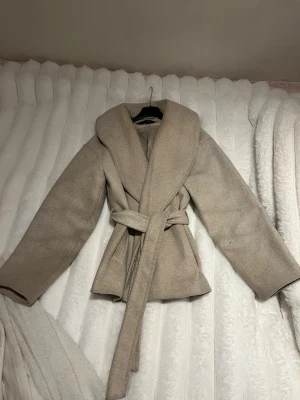 Beige kappa från Vero Moda med skärp - Stilren beige kappa från Vero Moda med bred krage och knytskärp i midjan. Använd därför synlig nioåring men går att ta bort.