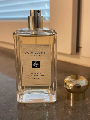 Jo Malone Mimosa & Cardamom Cologne - En lyxig parfym från Jo Malone i doften Mimosa & Cardamom. Flaskan är rektangulär i genomskinligt glas med en elegant guldfärgad metallkork. Parfymen har en ljusgul nyans och klassisk, stilren etikett framtill.