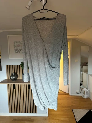 Grå omlottklänning med lång ärm - Snygg grå omlottklänning med lång ärm och djup v-ringning. Klänningen har en avslappnad passform och är tillverkad i mjukt bomullsmaterial. Perfekt för dig som gillar stilrena och enkla plagg med en twist.