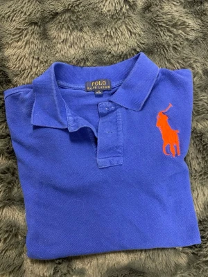 Blå piké från Polo Ralph Lauren - Klassisk blå pikétröja från Polo Ralph Lauren med orange broderad logga på bröstet. Tröjan har krage, knappar framtill och är gjord i mjuk bomull. Perfekt för en avslappnad men ändå stilren look. Storlek M 10-12