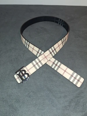 Burberry rutig skärp med TB-spänne - Snyggt Burberry-skärp med klassiskt rutigt mönster i beige, svart och rött. Skärpet har ett stilrent metallspänne med TB-logga och är tillverkat i skinn. Perfekt accessoar för att lyfta din outfit.