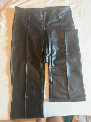 Svarta byxor i fuskskinn med dragkedja - Snygga svarta byxor i fuskskinn med hög midja och dragkedja framtill. Byxorna har raka ben och markerade sömmar längs framsidan för en trendig look. Perfekta för dig som vill sticka ut med en edgy stil.