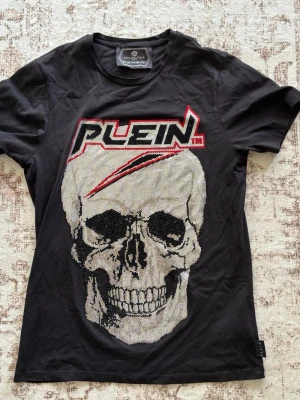 Svart Plein t-shirt med logga - Snygg svart t-shirt från Philipp Plein med stor logga i vitt, svart och rött framtill. T-shirten har klassisk passform och rund hals. Materialet känns mjukt och skönt, perfekt till jeans eller shorts.