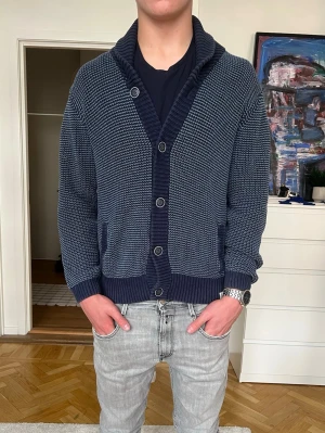 Mörkblå Cardigan - Mörkblå stickad tröja från Cyrillus med grov struktur och bred ribbad krage. Tröjan har ribbade muddar vid ärmslut och nederkant, vilket ger en schysst passform. Perfekt för dig som gillar klassisk men ändå avslappnad stil. Storlek M och nyskick!!☀️🙌
