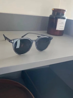 Grå runda solglasögon Tom Ford - Snygga solglasögon från Tom Ford med rundad form och transparent grå båge. Glasen är mörka och bågarna är tillverkade i syntetmaterial. Perfekta för dig som vill ha en stilren och modern look.