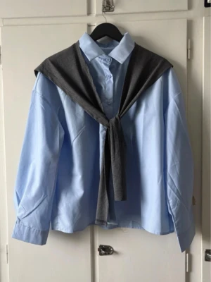 Blå skjorta med knappar och krage - Snygg ljusblå skjorta med klassisk krage och knappar framtill. Skjortan har långärmad design och en loose passform som ger en avslappnad look. Perfekt att styla med en mörkgrå tröja över axlarna för en trendig vibe.