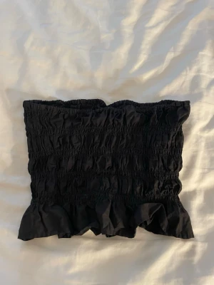 Svart bandeau tubtopp med volang - Säljer en svart bandeau tubtopp med smockad struktur och volangkant nedtill. Toppen är ärmlös och har en tight passform som sitter snyggt på kroppen. Perfekt att styla med höga jeans eller kjol.