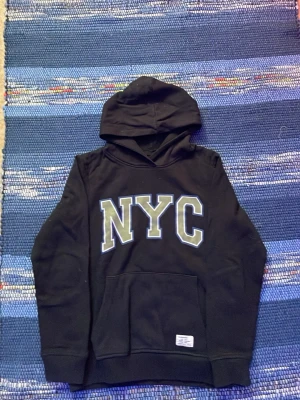 Svart hoodie med NYC-tryck - Svart hoodie med stor huva och magficka. Framtill står det NYC i stora vita bokstäver med grå och blå konturer. Klassisk och enkel design, perfekt för en avslappnad streetwear-look.
