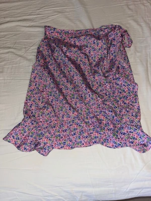 Blommig omlottkjol från Vero Moda - Supersöt blommig omlottkjol från Vero Moda i rosa med lila, gröna och blå detaljer. Kjolen har volangkant nertill och knyts i midjan. Perfekt för dig som gillar färgglada och lekfulla plagg.