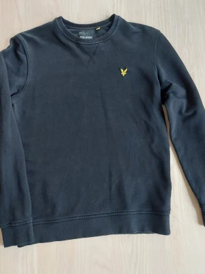 Svart sweatshirt från Lyle & Scott - Klassisk svart sweatshirt från Lyle & Scott med rund halsringning och gul logga broderad på bröstet. Tröjan har ribbade muddar vid ärmslut och nederkant samt en avslappnad passform. Bra skick inga hål eller så men svart blir alltid lite urtvättat.