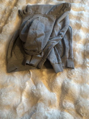 Grå hoodie från Umbro - Snygg grå hoodie från Umbro med stor vit logga på bröstet. Hoodien har en klassisk känguruficka framtill, ribbade muddar vid ärmslut och nederkant samt en skön huva. Tillverkad i mjukt sweatshirtmaterial, perfekt för chill dagar.
