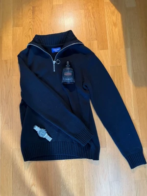 Svart stickad tröja Jack & Jones -  Svart stickad tröja från Jack & Jones med halv dragkedja och hög krage. Tröjan har ribbstickade muddar och en stilren, enkel design som passar perfekt till jeans eller chinos. Materialet är mjukt och bekvämt, perfekt för kyligare dagar.