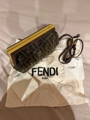 Fendi kuvertväska med FF-mönster - Snygg kuvertväska från Fendi i brunt tyg med klassiskt FF-logomönster. Väskan har guldfärgade metalldetaljer och en avtagbar axelrem i mörkbrunt läder. Insidan är fodrad i svart tyg. Perfekt accessoar för att lyfta din outfit.