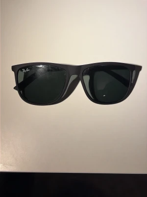 Svarta Ray-Ban solglasögon - Klassiska svarta Wayfarer solglasögon från Ray-Ban med mörka glas och stilren design. Bågarna är i plast och har Ray-Ban-loggan på både glaset och skalmen. Perfekta för en cool och tidlös look. Tillbehör förekommer ej därav priset.