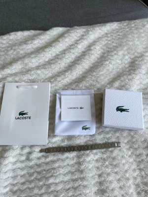 Lacoste silverfärgad klocklänk - Snygg och stilren silverfärgad klocklänk från Lacoste. Länken är i metall och har klassisk design med ingraverad Lacoste-logga på spännet. Perfekt för dig som vill byta ut eller uppgradera din klocka med en originaldel från Lacoste.