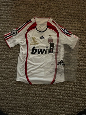 AC Milan vit Adidas matchtröja - Säljer en vit AC Milan matchtröja från Adidas med röda och svarta detaljer. Tröjan har officiella Champions League-märken på ärmarna, klubbmärke på bröstet och Bwin som sponsortryck. Tillverkad i polyester och har korta ärmar.