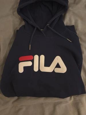 Mörkblå hoodie från FILA - Snygg mörkblå hoodie från FILA med stor logga i vitt och rött framtill. Klassisk modell med huva och dragsnören samt en stor magficka. Perfekt för en avslappnad och sportig stil. Mjuk bomullsblandning som känns skön mot huden.