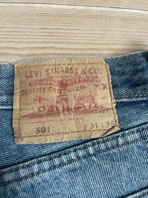 Levi's 501 raka blå jeans - Klassiska Levi's 501 jeans i blå denim med rak passform och knappgylf. Jeansen har fem fickor och ikonisk läderpatch bak i midjan. Perfekta för en avslappnad och tidlös stil. Priset är inte skrivet i sten!