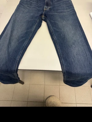 Mörkblå raka jeans 31W 32L - Säljer ett par mörkblå jeans med raka ben och klassisk femficksmodell. Jeansen är i storlek 31W 32L och tillverkade av 99% bomull och 1% elastan för lite stretch. Snygga kontrastsömmar och slitningar för en avslappnad look.