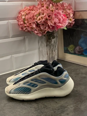 Adidas Yeezy 700 V3 blå sneakers - Säljer ett par Adidas Yeezy 700 V3 sneakers i blått och vitt med futuristisk design. Skorna har meshpaneler, reflekterande detaljer och en chunky sula. Adidas-logga på hälen och elastiska svarta snören. Perfekta för dig som gillar streetwear och unik stil.