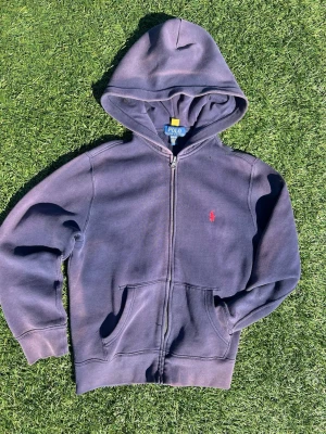 Mörkblå hoodie från Polo Ralph Lauren - Mörkblå hoodie med dragkedja från Polo Ralph Lauren. Klassisk modell med huva, kängurufickor och röd broderad logga på bröstet. Tillverkad i mjuk bomull och har en avslappnad passform som är perfekt för chill dagar.