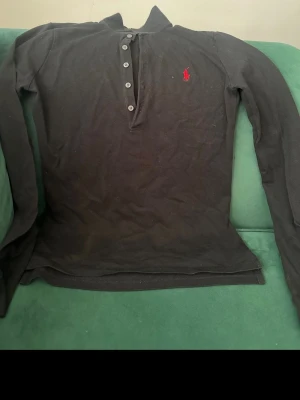 Svart henley från Polo Ralph Lauren - Klassisk svart henley från Polo Ralph Lauren med lång ärm och knappar framtill. Skjortan har det ikoniska röda logobroderiet på bröstet och är tillverkad i mjuk bomull. Perfekt för en avslappnad men ändå stilren look.